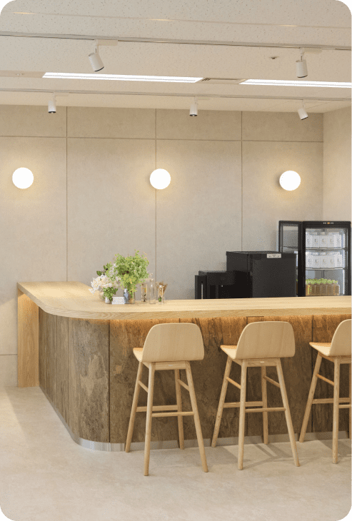ナチュラルなデザインのオフィスカウンター。木製のカウンターとスツールが並び、壁には丸い照明が配置されている。